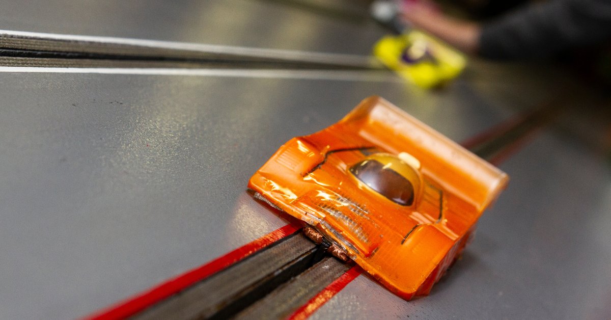 A close up of an orange slot car driving down a slot car track with a blurry yellow slot car behind it.